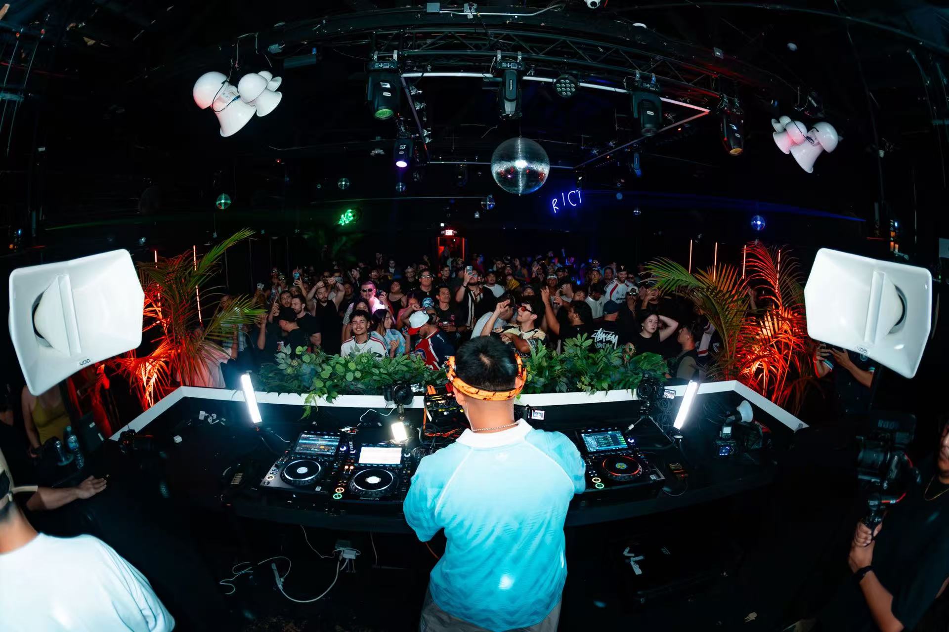 View from behind a DJ in a light blue shirt facing the dance-floor crowd.