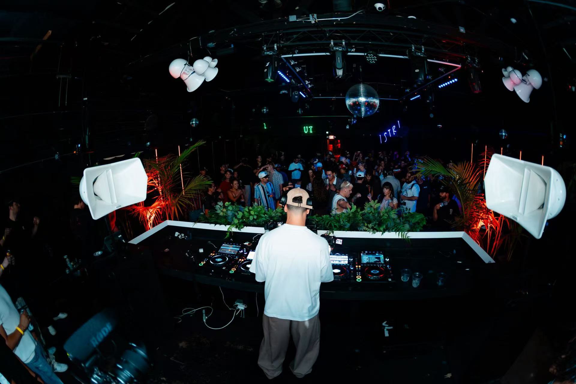 A DJ in a white shirt at the decks, facing a crowd beneath overhead lighting.