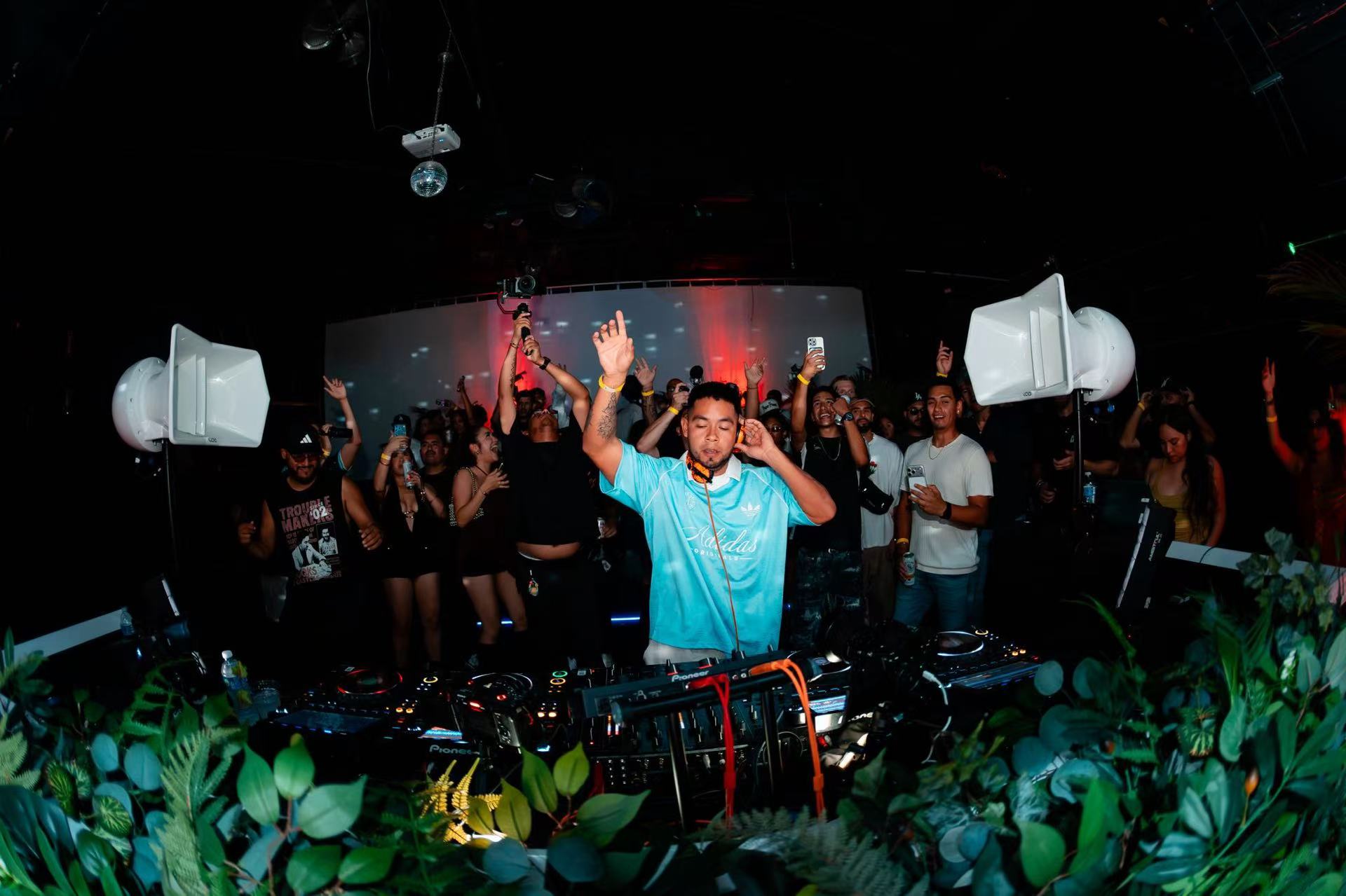 A DJ raising one hand behind a booth lined with foliage, with guests gathered behind.