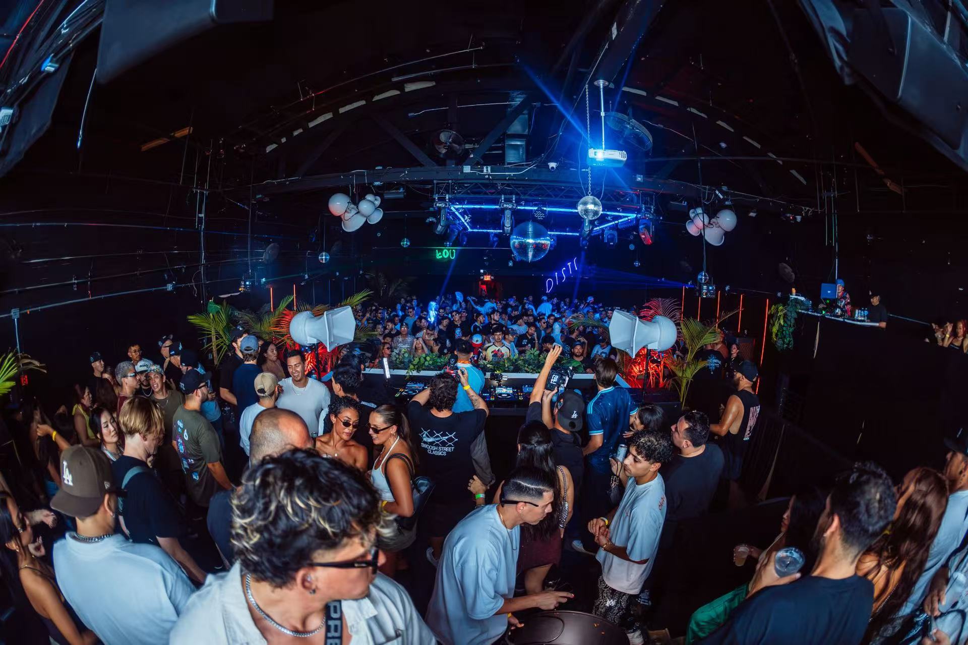 Wide view of a crowded dance floor beneath an exposed ceiling and blue lighting.